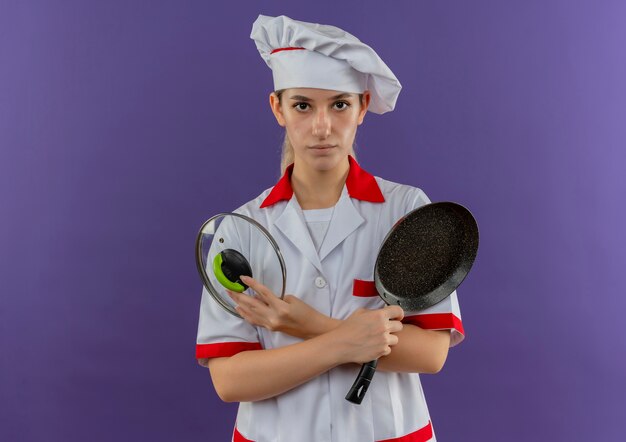 프라이팬과 팬 뚜껑을 들고 요리사 유니폼을 입은 젊은 예쁜 요리사