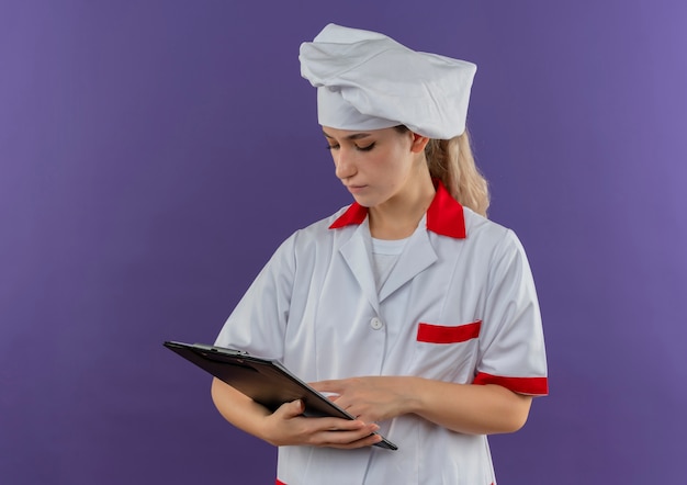 Young pretty cook in chef uniform holding looking at and putting finger on clipboard 
