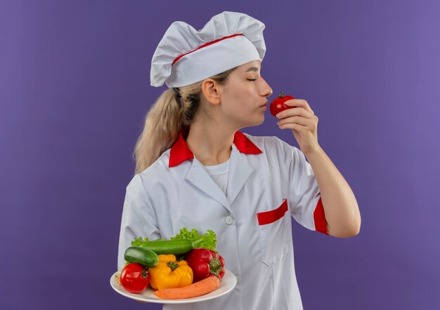 Молодой симпатичный повар в униформе шеф-повара держит тарелку с овощами и нюхает помидоры