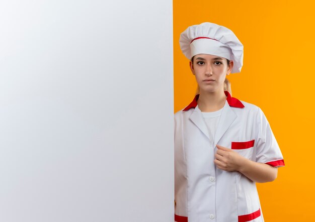 Young pretty cook in chef uniform standing behind white wall putting hand on her uniform looking 