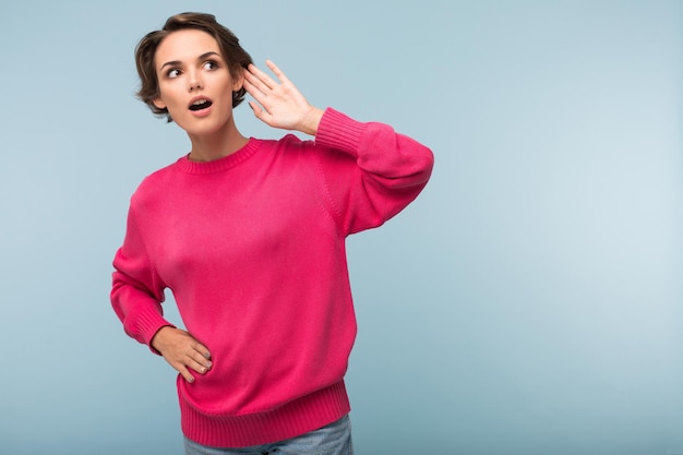 Young pretty curious woman with dark short hair in pink sweater holding hand over ear trying to listen something while amazedly looking aside over blue background