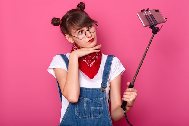 Young pretty dark haired women taking selfie on cell phone, using selfie stick, keeps hands under chin