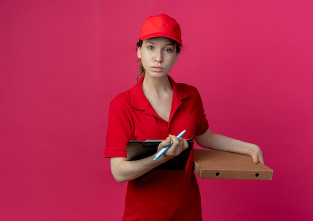 Молодая красивая доставщица в красной форме и кепке держит ручку пакета пиццы и буфер обмена, глядя в камеру, изолированную на малиновом фоне с копией пространства