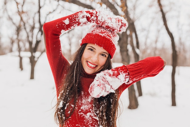 Giovane donna felice sorridente candida piuttosto eccitata in guanti rossi e cappello che indossa maglione lavorato a maglia a piedi giocando nel parco nella neve, vestiti caldi, divertendosi