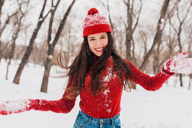雪の中で公園で遊んで、暖かい服を着て、楽しんで、赤いミトンとニットのセーターを着て歩いている帽子をかぶった若いかなり興奮した率直な笑顔の幸せな女性