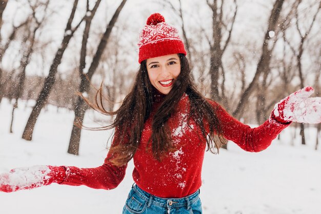 Young pretty excited candid smiling happy woman in red mittens and hat wearing knitted sweater walking playing in park in snow, warm clothes, having fun