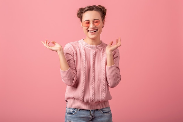 Молодая довольно возбужденная искренне смеющаяся женщина в розовом свитере и солнцезащитных очках изолирована на розовом студийном фоне, откровенная улыбка