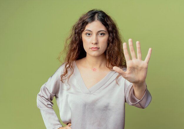 молодая симпатичная женщина-офисный работник показывает жест стоп и кладет руку на бедро, изолированную на оливково-зеленой стене