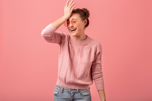 Young pretty funny emotional laughing woman in pink sweater and sunglasses isolated on pink studio background