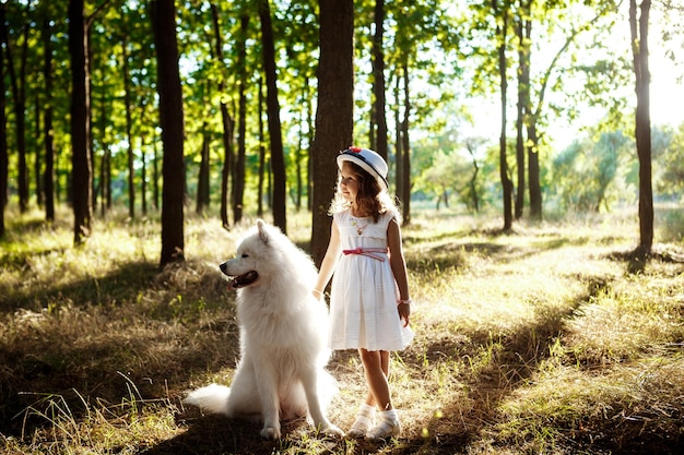 Young pretty girl in dress and hat walking, playing with white dog in park at sunset.