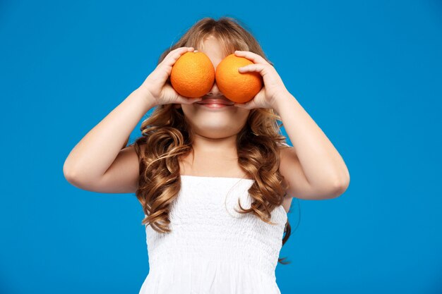 Молодая красивая девушка скрывает глаза остроумие апельсины над синей стеной
