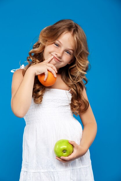 Молодая милая девушка держа яблоко и апельсин над голубой стеной