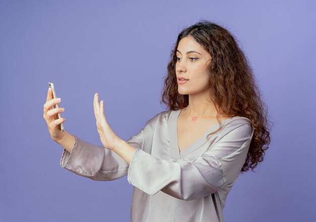 Молодая красивая девушка держит и смотрит в телефон и показывает жест остановки, изолированный на синем