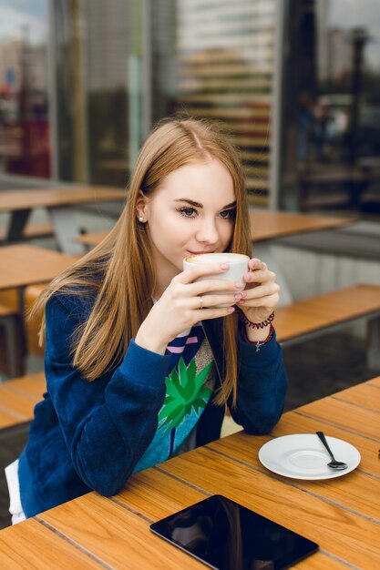 若い可愛い女の子がカフェの外のテーブルでコーヒーを飲んでいます。彼女は長い髪と青いジャケットを持っています。