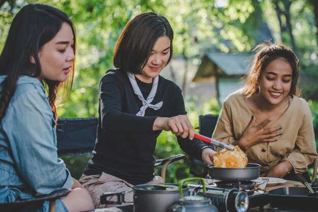 어린 예쁜 소녀는 자연 공원에서 친구들과 함께 캠핑을 하는 동안 냄비에 계란을 올려 놓고 간단한 식사를 요리하며 함께 토론하고 즐겁게 웃고 있습니다.