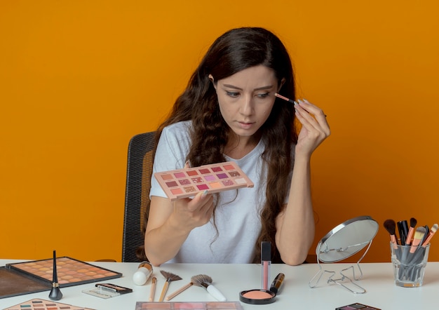 無料写真 ミラーを見て、オレンジ色の背景に分離されたアイシャドウを適用する化粧ツールで化粧テーブルに座っている若いかわいい女の子