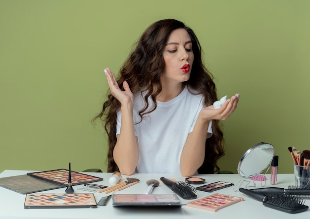Young pretty girl sitting at makeup table with makeup tools holding looking and blowing at hair mousse and keeping another hand in air isolated on olive green background