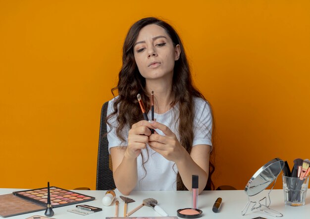 Young pretty girl sitting at makeup table with makeup tools holding and looking at concealer and eyeshadow brushes isolated on orange background