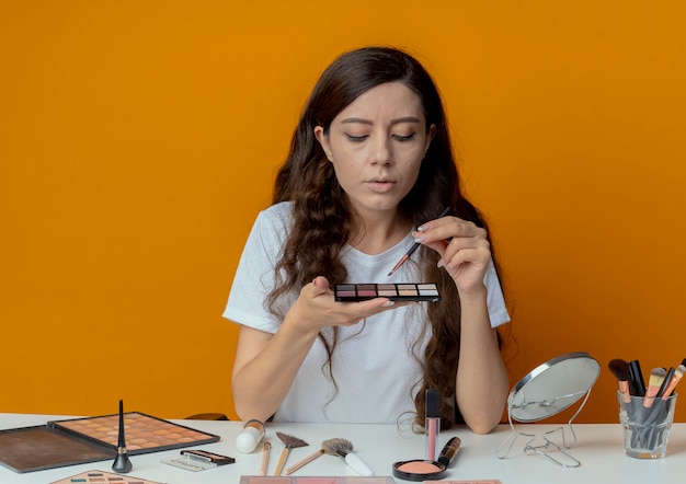 Young pretty girl sitting at makeup table with makeup tools holding and looking at eyeshadow palette and holding eyeshadow brush isolated on orange background