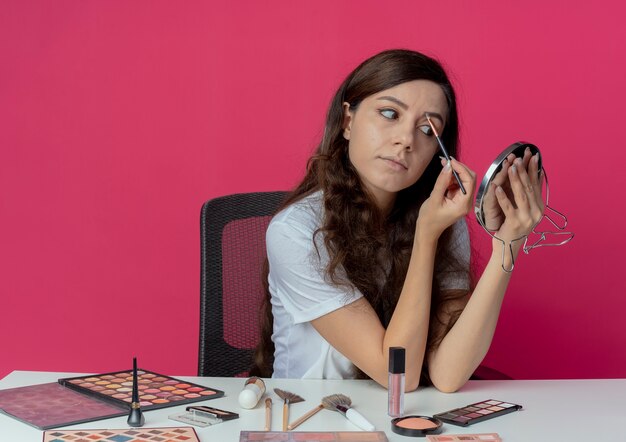 Young pretty girl sitting at makeup table with makeup tools holding and looking at mirror shaping her eyebrow with makeup brush isolated on crimson background