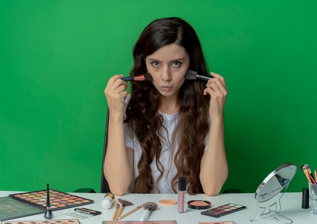 Young pretty girl sitting at makeup table with makeup tools holding powder and blush brushes and touching cheeks with them and looking at camera isolated on green background