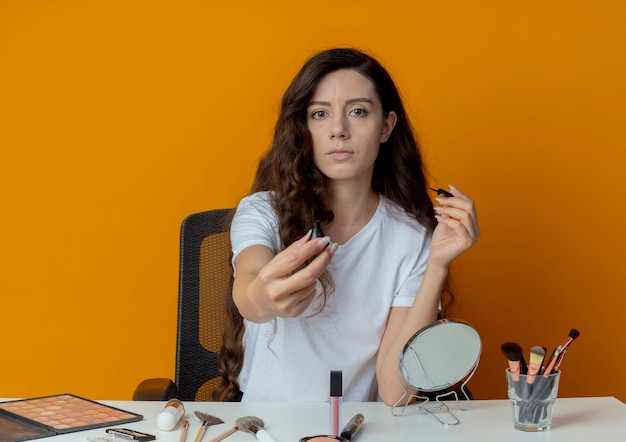 Young pretty girl sitting at makeup table with makeup tools holding and stretching out eyeliner towards camera and looking at camera isolated on orange background