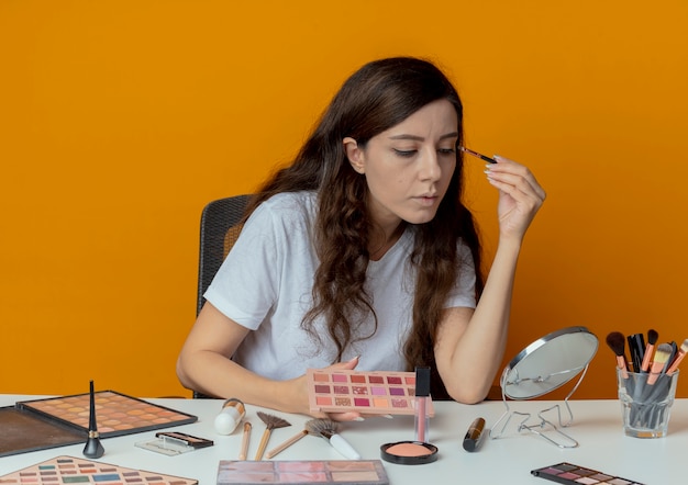 ミラーを見て、オレンジ色の背景に分離されたアイシャドウを適用する化粧ツールで化粧テーブルに座っている若いかわいい女の子