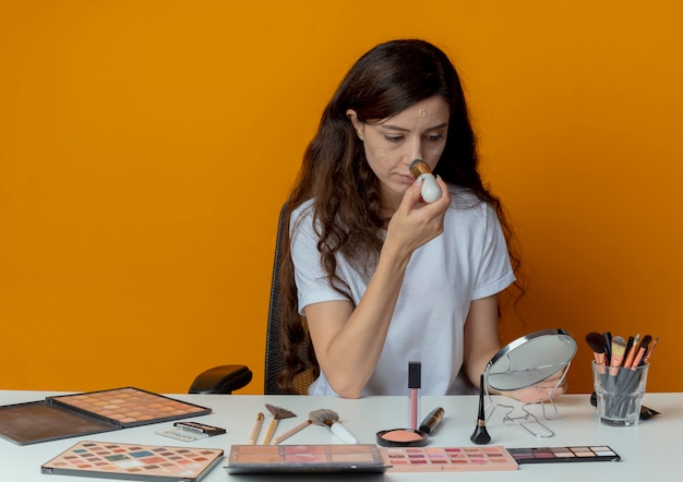 Young pretty girl sitting at makeup table with makeup tools looking at mirror and applying foundation isolated on orange background