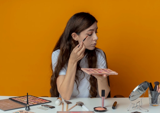 Young pretty girl sitting at makeup table with makeup tools looking at mirror holding shadow palette and applying eyeshadow with brush isolated on orange background