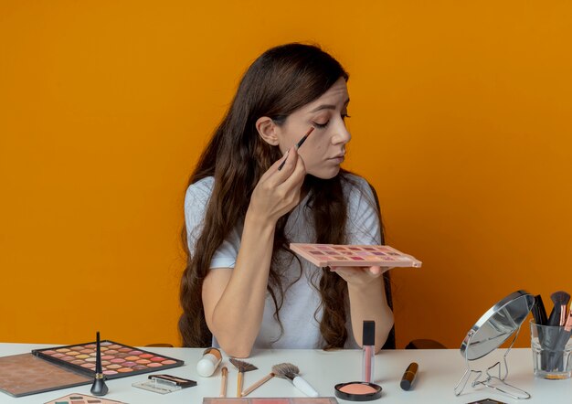 Молодая красивая девушка сидит за косметическим столом с инструментами для макияжа, глядя в зеркало, держа палитру теней и применяя тени для век кистью, изолированной на оранжевом фоне