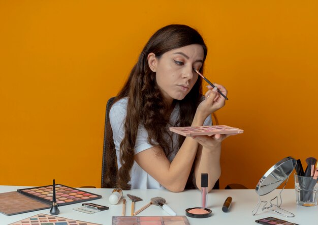 Young pretty girl sitting at makeup table with makeup tools looking at mirror holding shadow palette and applying eyeshadow with brush isolated on orange background