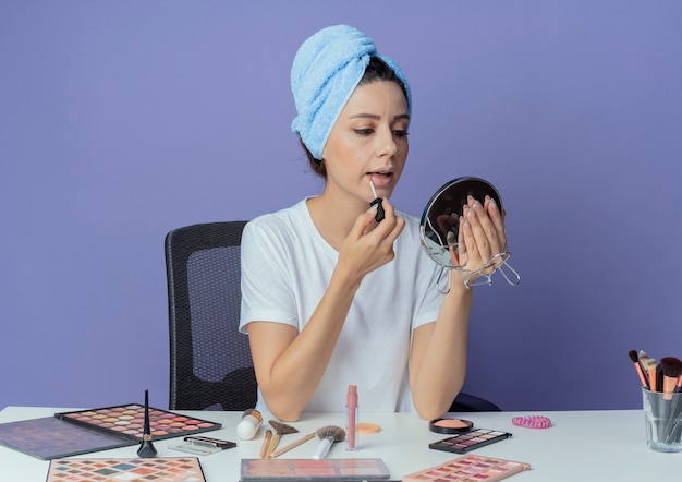 Young pretty girl sitting at makeup table with makeup tools and with bath towel on head holding and looking at mirror and applying lipgloss isolated on purple background