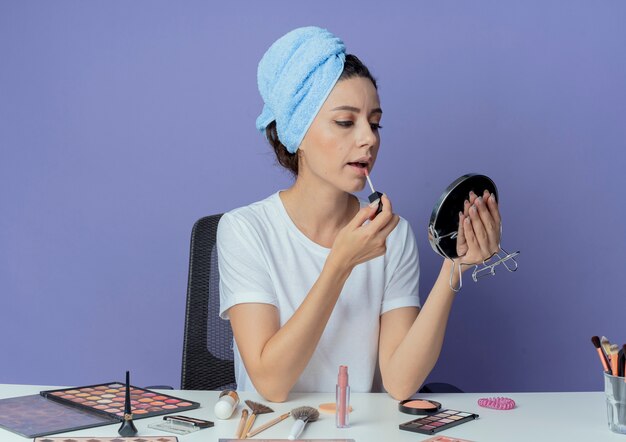 Young pretty girl sitting at makeup table with makeup tools and with bath towel on head holding and looking at mirror and applying lipgloss isolated on purple background