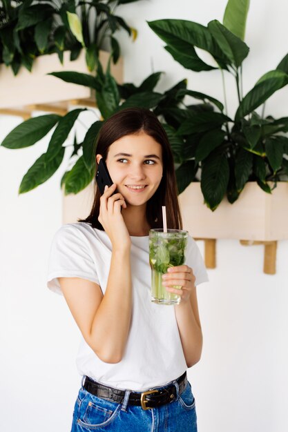 携帯電話を話していると、カフェのテーブルに立っている間スムージーを飲む若いきれいな女の子