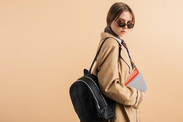 Free photo young pretty girl in trench coat and sunglasses with backpack on shoulder holding notepads in hand thoughtfully looking in camera while spending time over beige background