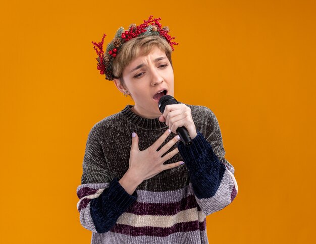 Young pretty girl wearing christmas head wreath holding microphone looking down keeping hand on chest singing isolated on orange wall with copy space