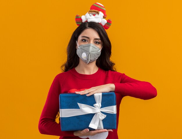 Young pretty girl wearing santa claus headband and sweater with protective mask  holding gift package isolated on orange wall