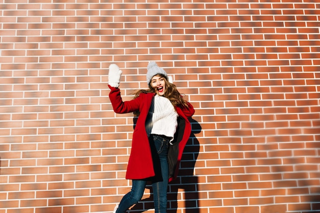 Young pretty girl with long hair in red coat, hat, gloves having fun  on wall  outside.