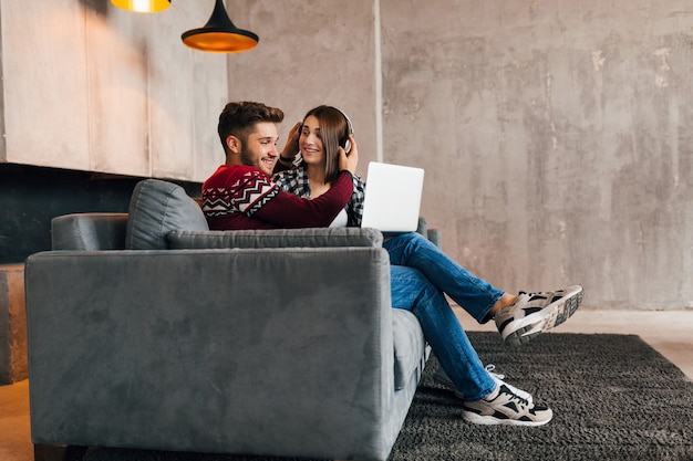 Young pretty happy smiling man and woman sitting at home in winter, looking in laptop, listening to headphones, students studying online, couple on leisure time together,