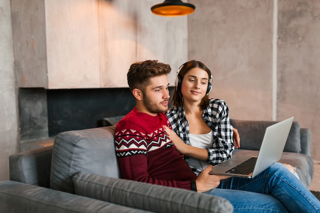 Young pretty happy smiling man and woman sitting at home in winter, looking in laptop, listening to headphones, students studying online, couple on leisure time together,