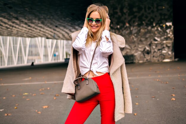 Young pretty hipster  woman posing on the street near modern business centers , wearing trendy office outfit and cashmere coat , sending kiss and enjoyed cool autumn day, toned colors.