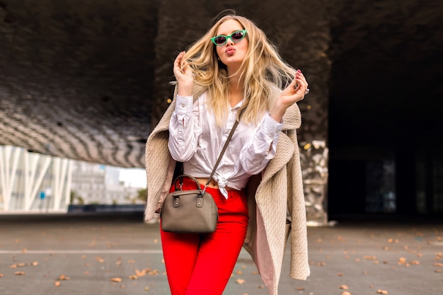 Young pretty hipster  woman posing on the street near modern business centers , wearing trendy office outfit and cashmere coat , sending kiss and enjoyed cool autumn day, toned colors.