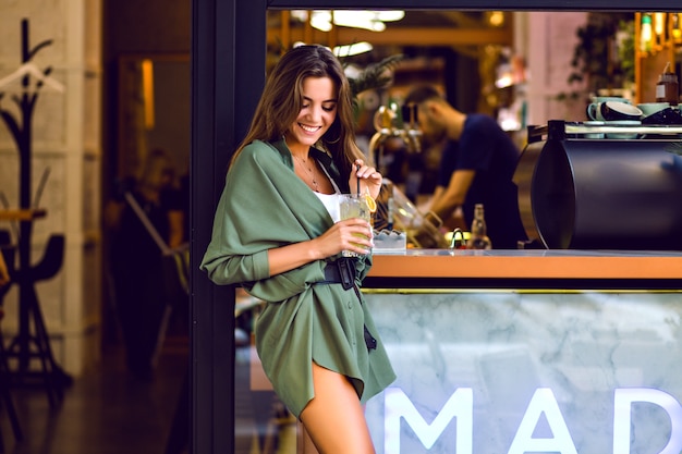 Young pretty lady enjoying free time at city cafeteria and bar, drinking lemon and having fun, trendy hipster outfit, toned colors.