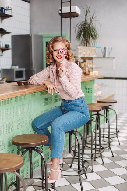 Giovane bella signora in camicia e jeans seduta al bancone del bar e coprendosi l'occhio con caramelle lecca-lecca mentre guarda con gioia in camera nella caffetteria