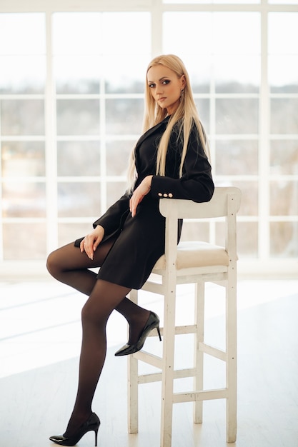 Free photo young pretty lady sitting on white chair while posing with window