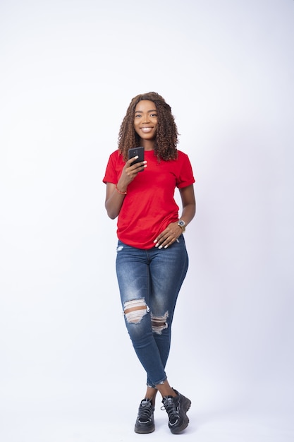 Young pretty lady standing with legs crossed holding her phone and smiling