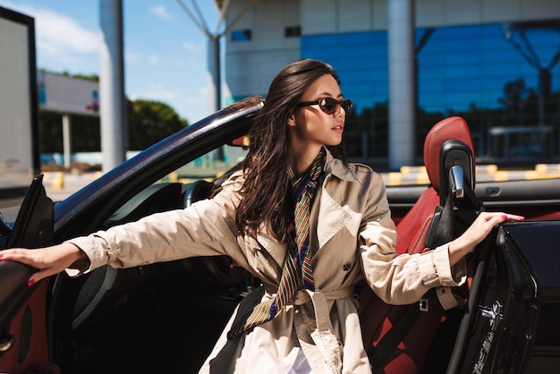 Free photo young pretty lady in sunglasses and trench coat opening cabriolet car door while dreamily looking aside with airport on background