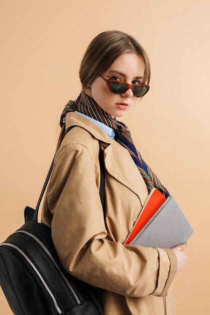 Young pretty lady in trench coat and sunglasses with backpack on shoulder holding notepads in hand while dreamily looking in camera over beige background