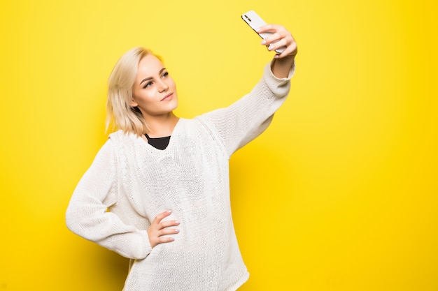 白いセーターの若いきれいな女性女性女の子は黄色の彼女のスマートフォンでselfieを作る