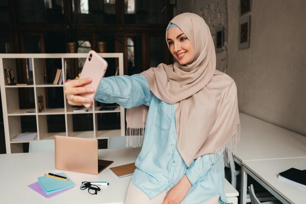 Young pretty modern muslim woman in hijab working on laptop in office room, education online